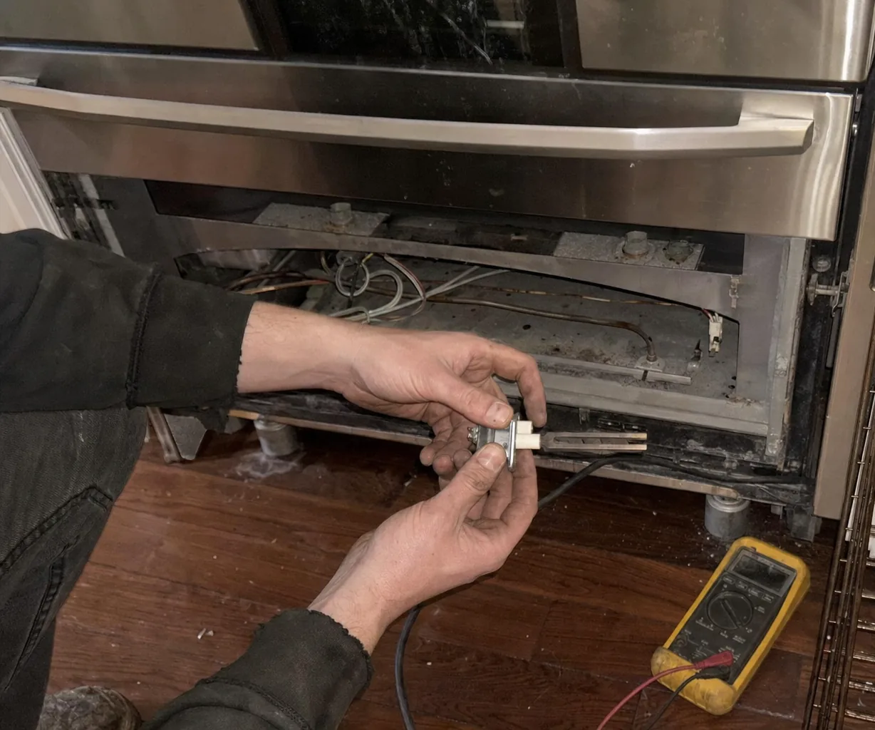 Lower front of a wide stainless range with its access panel off, burner tube and wiring loom visible above an old timber floor