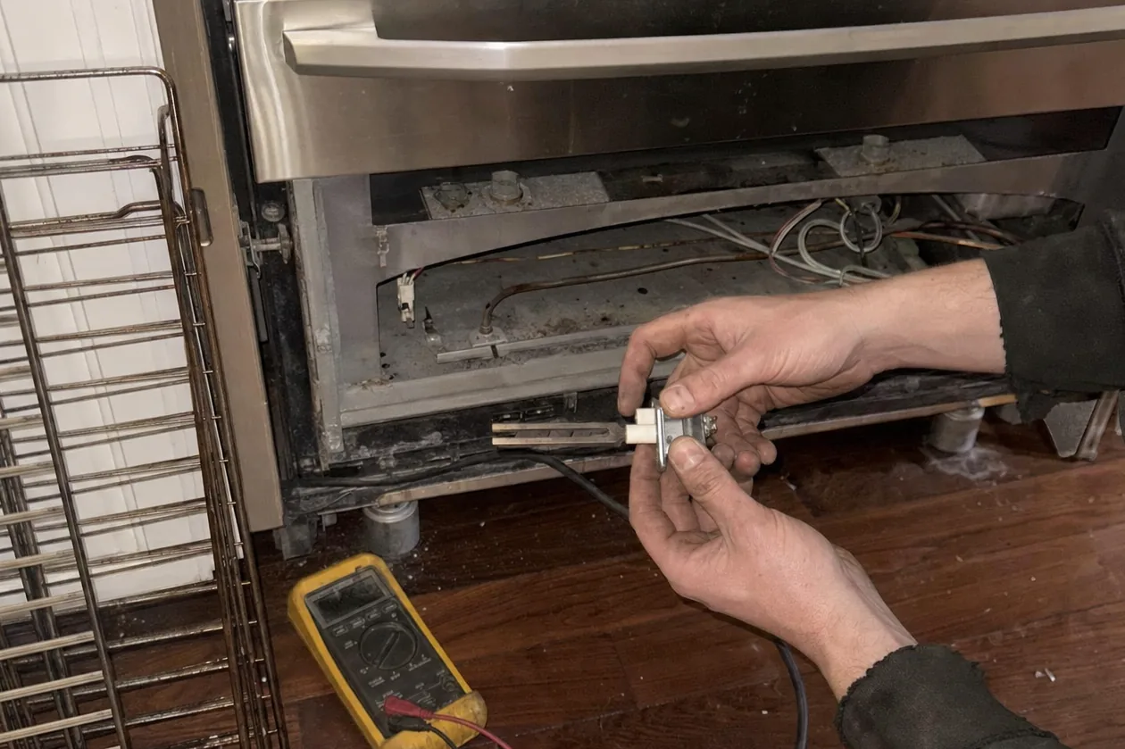 Two hands holding an oven igniter and its metal bracket clear of the open lower panel of a range, a yellow multimeter and a wire oven rack resting on the floorboards nearby