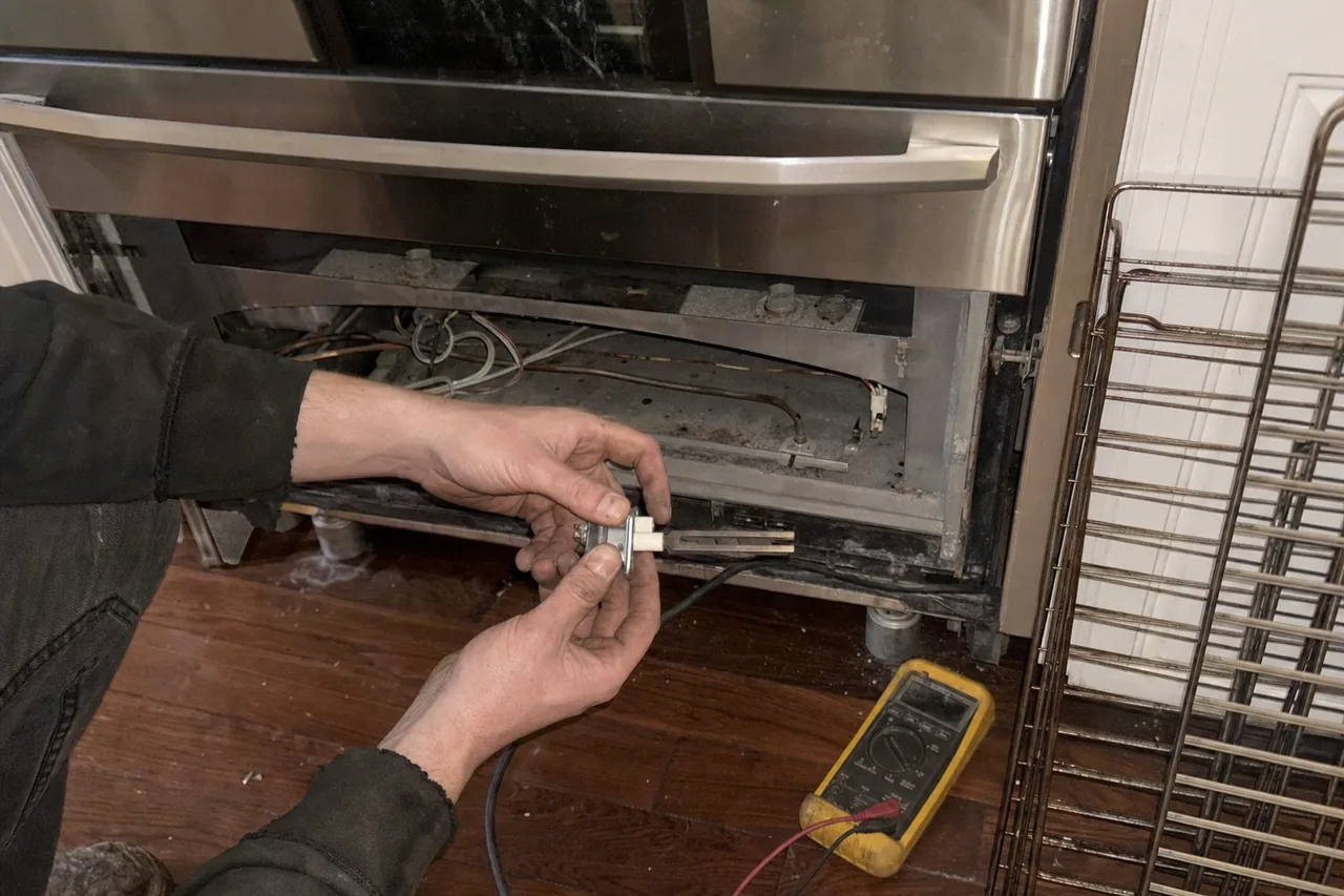 Technician kneeling at a stainless range with the lower panel removed, holding the oven igniter and its ceramic mount clear of the exposed wiring while a yellow multimeter lies on the floor
