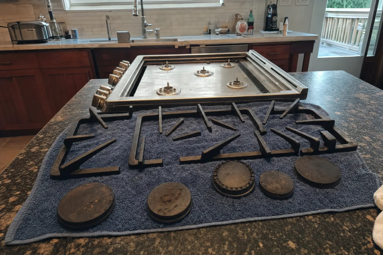 Gas cooktop stood on a granite island with all grates removed, showing the slotted downdraft vent running along its back edge