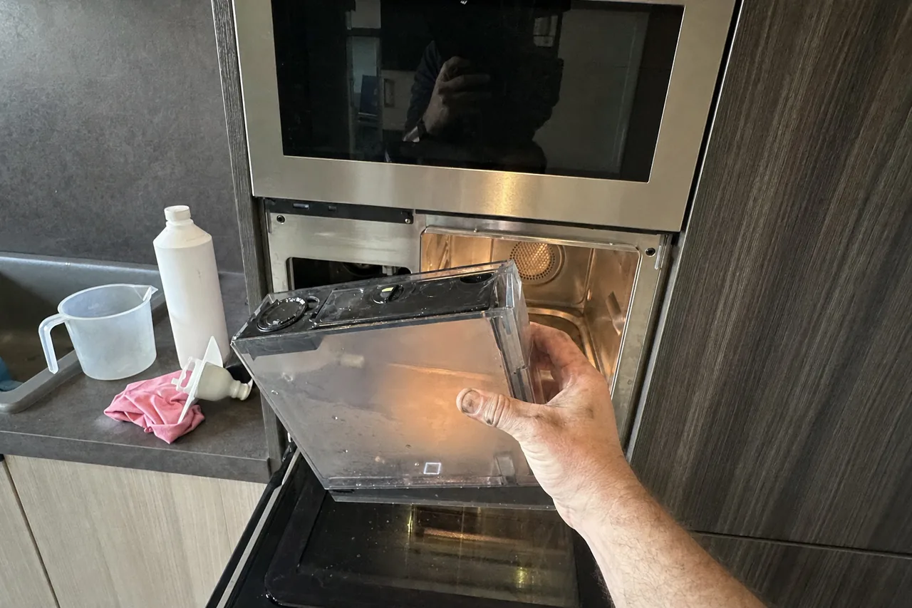 Clear water reservoir lifted out of a built-in steam oven set into a dark timber cabinet column, descaling bottle and measuring jug on the counter