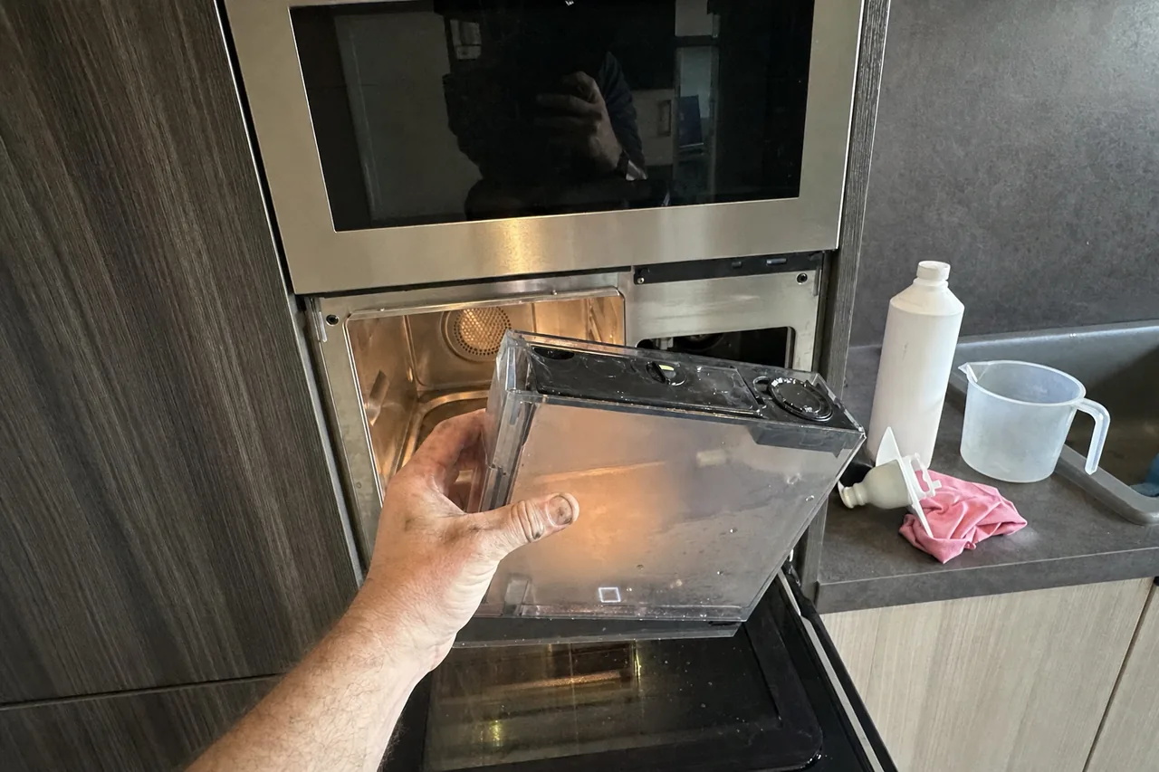 Technician lifting a clear water reservoir out of a built-in convection steam oven set into a dark cabinet column