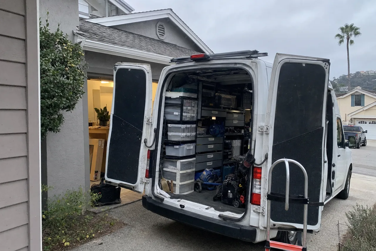 Service van standing on a wide driveway apron beside a house, rear doors open on shelves of labelled parts bins and a strapped hand truck