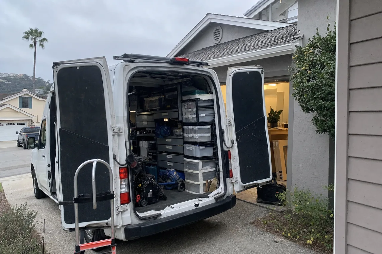 Service van with its rear doors open in front of an open garage, hand truck ready at the tailgate and parts shelving visible inside